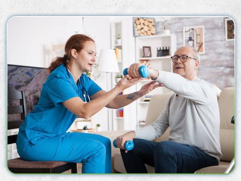 A female nurse in blue scrubs assists an elderly male patient with a dumbbell exercise focused on orthopedic health in a living room.